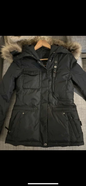 Svart pufferjacka med fuskpäls - Svart pufferjacka med huva och fuskpäls, flera fickor med både dragkedja och knappar. Jackan har lång ärm och quiltad design, perfekt för kalla dagar. Snygg och funktionell med detaljer som tryckknappar och dragkedja framtill.