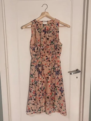 Blommig sommarklänning Dry Lake S - Superfin ärmlös klänning från Dry Lake i storlek S. Klänningen är ljusbeige/rosa med färgglatt blommigt mönster i massa olika färger! Klänningen har resår i midjan och är tillverkad i ett lätt, flowy material med innerfoder. Perfekt för varma dagar och somriga vibes! Den har en kedja fastsydd i efterhand stället för en vanlig knapp (se sista bilden), men jag tycker att det bara bidrar till sommarviben!🌸