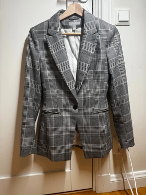 Grå rutig kavaj från H&M - Snygg grå kavaj från H&M med klassiskt rutigt mönster och en knapp framtill. Kavajen har skarpa axlar, smal passform och två fickor fram. Perfekt för dig som gillar stilrena och trendiga plagg med lite edge.