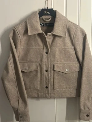 Beige overshirt från Zara - Snygg beige overshirt från Zara med klassisk krage och stora fickor framtill. Jackan har knappar längs hela framsidan och är tillverkad i mjukt ullmaterial. Perfekt för lager-på-lager och har en clean, modern look.