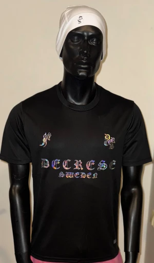Decrese Sweden – Black Gloss Edition - Decrese Sweden – Black Gloss Edition Svart T-shirt med holografiskt tryck som skiftar i färg för en sportig, modern streetwear-look. Lätt och bekväm med matchtröjekänsla. Perfekt till vardag, gym och event. Extra snygg ihop med vit Decrese-mössa.