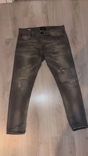 Grå slim fit jeans från Jack & Jones - Säljer ett par grå slim fit jeans från Jack & Jones med slitna detaljer och patch på ena benet. normal midja och är tillverkade i stretchig denim för en slim fit passform!