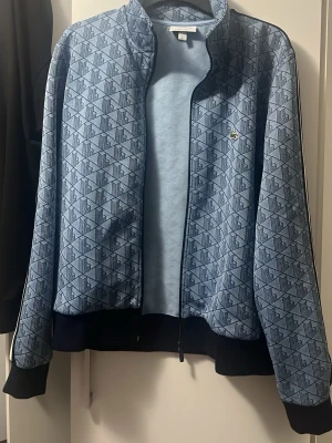 Blå mönstrad zip hoodie från Lacoste  - Säljer en blå hoodie från Lacoste med hel dragkedja och svart ribbade muddar. Tröjan har ett geometriskt mönster över hela ytan och den klassiska loggan på bröstet. Perfekt för dig som gillar sportig och stilren look.