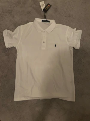 Vit pikétröja Polo Ralph Lauren S - Vit pikétröja från Polo Ralph Lauren i klassisk modell med krage och två knappar framtill. Tröjan har det ikoniska broderade logotypen på bröstet och är tillverkad i mjuk bomull. Perfekt för en clean och stilren look.