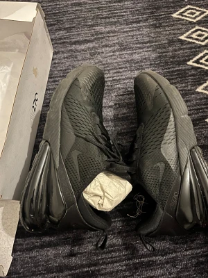 Nike Air Max 270 svarta sneakers - Nike Air Max 270 sneakers i helsvart färg med mesh-överdel och stor synlig Air-enhet i hälen.Den helt ny och inte använt , det finns alla storlekar,Snygg sportig design med snörning och platt sula, perfekt för dig som gillar streetstyle. Skorna har diskret Nike-logga på sidan och är tillverkade i syntet och mesh