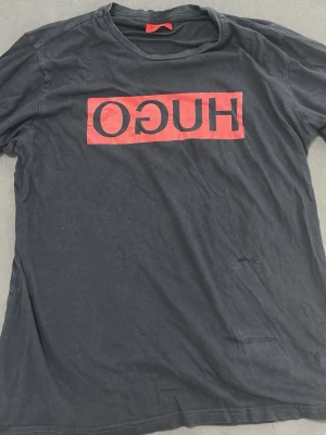 Svart HUGO t-shirt XXL med röd logga - Svart t-shirt från HUGO BOSS i storlek XXL med en fet röd logga på bröstet. Klassisk rund hals och korta ärmar. Tillverkad i mjuk bomull som känns skön mot huden. Perfekt för dig som gillar streetstyle och tydliga prints.