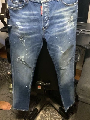 Blå Dsquared2 jeans med slitningar - Säljer ett par blå jeans från Dsquared2 med coola slitningar och färgstänk på framsidan. Jeansen har klassisk femficksdesign, låg midja och raka ben. Märkeslapp bak och Dsquared2-tag vid gylfen. Perfekt för dig som gillar streetstyle och detaljer.