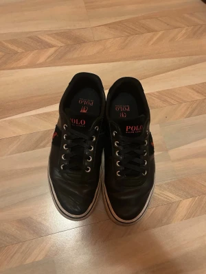 Svarta sneakers från Polo Ralph Lauren - Svarta sneakers från Polo Ralph Lauren med klassisk låg profil och rund tå. Skorna har snörning med metallöglor, vit platt sula och röd Polo-logga på sidan och innersulan. Materialet är skinn och textil, vilket ger en stilren och sportig vibe. Passar även 40 41 och 42 på bredden 