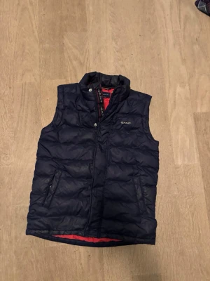 Mörkblå dunväst från GANT - Snygg mörkblå dunväst från GANT med röd insida och hög krage. Västen har två fickor med dragkedja och quiltad design. Perfekt för lager-på-lager och håller dig varm under kyliga dagar. Klassisk look med diskret logga på bröstet.