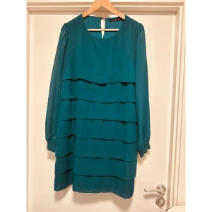 Turkos kortklänning från Atmosphere - Snygg turkos klänning från Atmosphere med långärmade transparenta ärmar och knäppning vid manschetten. Klänningen har flera vågade lager framtill som ger en cool struktur och är tillverkad i ett lätt polyestertyg. Rund halsringning och knapp i nacken.