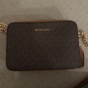 Brun axelväska Michael Kors.  - Säljer denna snygga  axelväskan från Michael Kors med MK-mönster, guldfärgade detaljer och en dekorativ tofs. Väskan är välanvänd men fortfarande i fint skick. Det finns slitage – guldfärgen på kedjan/dragkedjan har slitits något och på två ställen syns repor där tyget påverkats. 