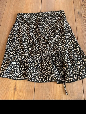 Leopardmönstrad kjol från SHEIN - Snygg kort kjol från SHEIN med leopardmönster i svart, beige och brunt. Kjolen har volangkant nertill och en liten knytning framtill. Perfekt för dig som vill sticka ut med ett djurmönstrat plagg.