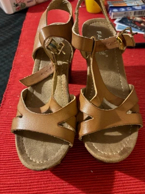 Bruna sandaler med klack och platå - Snygga bruna sandaler med bred klack och platåsula. Skorna har remmar i skinnimitation och justerbart spänne i guld. Perfekta för dig som vill ha lite höjd men ändå stabilitet. Passar till både jeans och klänning.