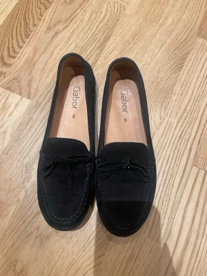 Svarta mockaloafers från Gabor size 37.5 - Snygga svarta loafers från Gabor i mjuk mocka med dekorativ rosett framtill och diskreta svarta pärlor. Skorna har rund tå och platt sula med bra grepp. Perfekt för dig som gillar klassisk stil med en twist. Använd 1 gång