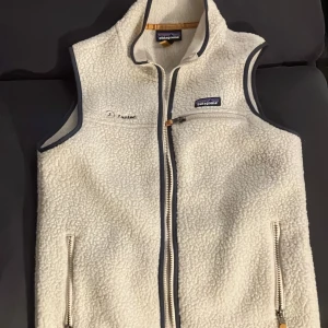 Vit fleeceväst från Patagonia M - Säljer en vit fleeceväst från Patagonia i storlek M. Västen har en mjuk och fluffig yta, hel dragkedja framtill, tre fickor med dragkedja och mörkgrå kantband. Perfekt för lager-på-lager och outdoor-häng. Perfekt skick helt o andvänd 