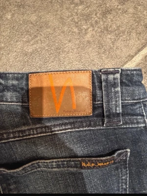 Blå slim fit jeans från Nudie Jeans - Snygga blå jeans från Nudie Jeans med klassisk läderpatch och orange logga bak. Modellen har smal passform och raka ben, med slitningar och tvättade detaljer för en cool look. Jeansen har fem fickor och är tillverkade i jeansmaterial. Längd 104 cm bredd 36 cm