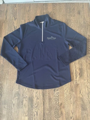 Marinblå golftröjor från Next Tee - Marinblå långärmad tröja från Next Tee Golf Clothes med vit half zip framtill. Tröjan har en stilren design med tryckt logga på bröstet och är gjord i ett lätt, syntetiskt material som passar perfekt för golf eller träning.