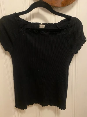 Svart ribbad topp från Lindex - Svart ribbad topp från Lindex med korta ärmar och söt volangkant vid halsringning, ärmslut och nederkant. Materialet är mjukt och stretchigt, perfekt för dig som gillar enkel men snygg stil. Passar dig som vill ha en basic topp med lite extra detalj.