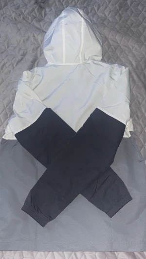 Under Armour vit och grå vindjacka - Snygg vindjacka från Under Armour i vitt och grått med svarta ärmar. Jackan har huva med meshfoder, dragkedja framtill och två sidofickor. Under Armour-logga i rosa på bröstet ger en sportig vibe. Perfekt för dig som gillar stilren och funktionell design.