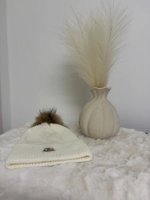 Vit  mössa med pälsboll - Ribbstickad vit mössa med stor brun pälsboll på toppen.. Mössan är mjuk och stretchig, perfekt för kalla dagar. Snygg och enkel design som ger en clean look.