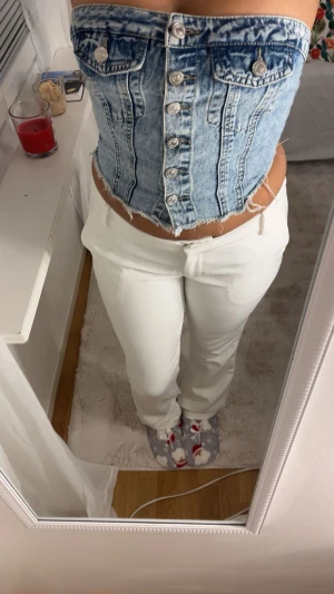 Blå jeanskorsett med knappar - Säljer en snygg blå jeanskorsett med silverfärgade knappar framtill och två dekorativa fickor. Korsetten har en croppad passform och rå kant nedtill för en cool vibe. Perfekt att styla med höga byxor eller kjol.