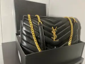 Snygg svart axelväska från Saint Laurent med quiltad design och guldiga kedjor. Väskan har YSL-logga i guld på framsidan och är tillverkad i skinn. Perfekt storlek för det viktigaste och ger en lyxig vibe till din outfit.