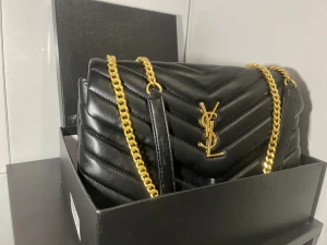   Saint Laurent axelväska - Snygg svart axelväska från Saint Laurent med quiltad design och guldiga kedjor. Väskan har YSL-logga i guld på framsidan och är tillverkad i skinn. Perfekt storlek för det viktigaste och ger en lyxig vibe till din outfit.