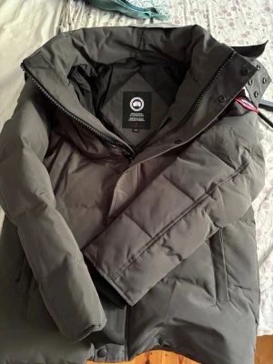 Grå Canada Goose Wyndham Parka - Säljer en grå Wyndham Parka från Canada Goose med ikonisk röd och vit patch på ärmen. Jackan har dunfyllning, huva och dragkedja framtill. Perfekt för kalla vinterdagar och har en clean, stilren look som funkar till allt. Medföljer originalpåse och etiketter.