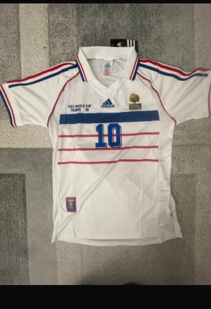 Adidas Frankrike Zidane VM 98 tröja - Säljer en ikonisk vit Frankrike landslagströja från VM 1998 med Zidane och nummer 10 på ryggen. Tröjan har blå och röda ränder, broderade detaljer och officiella FFF-märken. Perfekt för dig som älskar fotbollshistoria och retrostil.