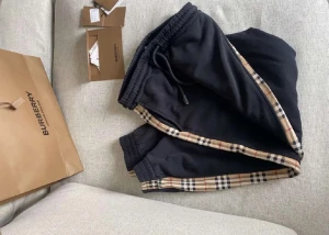 Svarta Burberry mjukisbyxor med rutiga detaljer - Svarta mjukisbyxor från Burberry med klassiska rutiga sidopaneler i beige, svart och rött. Byxorna har resår och snörning i midjan samt muddar vid bensluten. Perfekta för en avslappnad och trendig look.