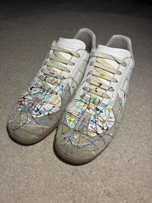 Maison Margiela sneakers med färgstänk - Unika vita sneakers från Maison Margiela med coola färgstänk i blått, rött, gult, grönt och svart. Skorna har beige mockadetaljer, snörning och platt gummisula. Perfekt för dig som vill sticka ut med en konstnärlig vibe och streetstyle.