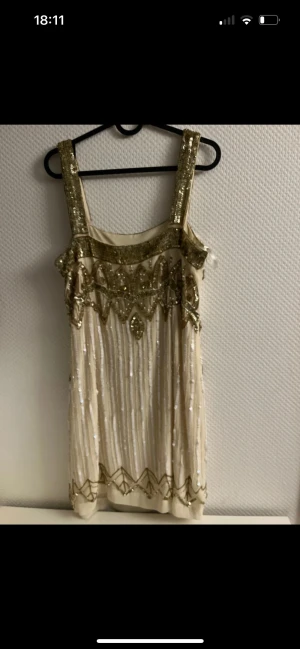 Glittrig kortklänning med paljetter - Superfin kortklänning i ljus champagnefärg med breda axelband och massor av guldiga paljetter och pärlor. Klänningen har ett glittrigt mönster med detaljer i guld och vitt, och är perfekt för dig som vill sticka ut. Materialet känns lyxigt och skimrande.