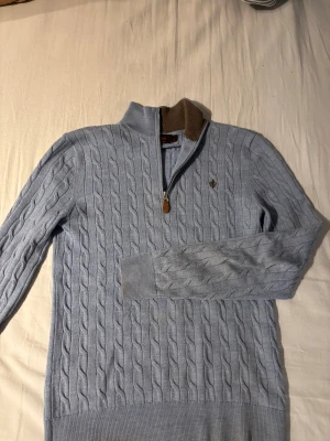 Ljusblå kabelstickad tröja Morris - Snygg ljusblå kabelstickad tröja från Morris i 100% merinoull. Den är storlek S men är ganska liten i storlek så den passar även XS. Tröjan har half zip med brunt foder i kragen och ett broderat emblem på bröstet. 