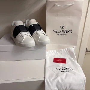 Valentino Garavani Open sneakers vit/svart - Snygga Valentino Garavani Open sneakers i vitt skinn med bred svart rem över vristen och coola nitar bak på sulan. Klassisk rund tå, vita snören och platt sula. Kommer med originalkartong och dustbag. Perfekta för dig som gillar lyxig streetstyle.