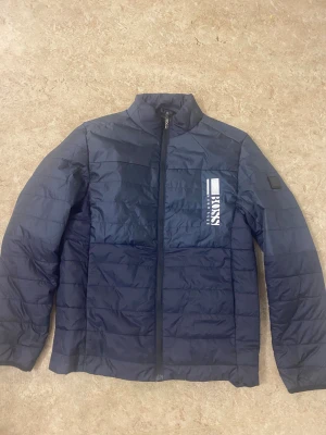 Mörkblå pufferjacka från Hugo Boss - Snygg mörkblå pufferjacka från Hugo Boss med vit logga på bröstet. Jackan har quiltad design, hög krage och hel dragkedja framtill. Tillverkad i syntetmaterial och har långärmad passform. Perfekt för dig som vill ha en stilren och modern look.