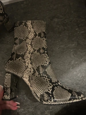 Ormskinnsmönstrade boots med klack - Säljer ett par boots med ormskinnsmönster i beige, svart och grå toner. Skorna har spetsig tå, blockklack och dragkedja på sidan. Materialet är syntet med reptilstruktur som ger en edgy vibe. Perfekt för dig som vill sticka ut med coola detaljer. orginalpris 800:-kr säljs endast för 499:-kr pris kan diskuteras. 🤎