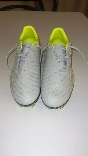 Nike Phantom GX fotbollsskor grå/gul - Nike Phantom GX PRO fotbollsskor med snygg grå ovandel och neon-gula detaljer. Skorna har snörning, rund tå och platt sula med dobbar för gräsplan och konstgräs. Användt de En gång, man kan även se en liten svart linje på bild tre, men man tänker knappt på det. Säljer de pga att de inte passa 