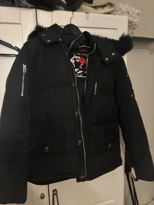 Svart dunjacka från Moose Knuckles - Svart dunjacka från Moose Knuckles äkta  med pälskantad huva och flera dragkedjor. Jackan har stora fickor framtill, tryckknappar och en broderad logga på insidan. Perfekt för kalla dagar och har en klassisk, stilren look.
