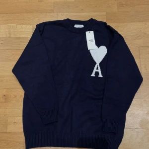 Helt ny stickad tröja från Ami - Snygg marinblå stickad tröja från Ami Alexandre Mattiussi med vit hjärtapplikation och bokstaven A på bröstet. Rund hals och ribbade muddar vid ärmslut och nederkant. Perfekt för dig som gillar stilrena detaljer och vill ha något som sticker ut.