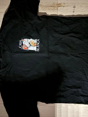 Svart Casa Blanca t-shirt med tryck - Svart t-shirt från Casa Blanca med stort färgglatt tryck på ryggen och mindre tryck på bröstet. Motivet har konstnärliga detaljer som palett, kolonner och frukt. Rund hals och korta ärmar, tillverkad i mjuk bomull. Perfekt för dig som gillar unika prints.