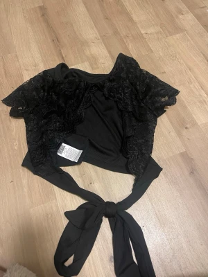 Svart spetsig bolerotopp från Firerie - Helt ny. Supersnygg svart bolerotopp med spetsdetaljer och öppen rygg. Toppen har korta ärmar i spets, ribbad framsida och knytband i midjan. Perfekt för att lyfta vilken outfit som helst med sin unika design och feminina vibe.