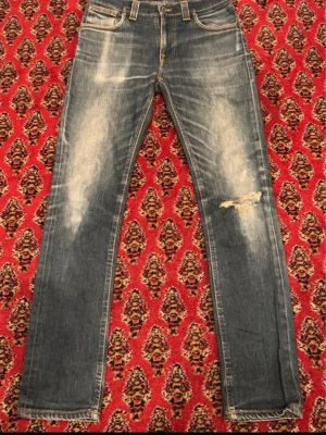 Blå Nudie Jeans med slitningar - Snygga blå jeans från Nudie Jeans med klassisk femficksdesign och coola slitningar på framsidan. Jeansen har kontrastsömmar, broderade bakfickor och en patch i skinn vid linningen. Modellen är rak och har en normal passform med låg midja.