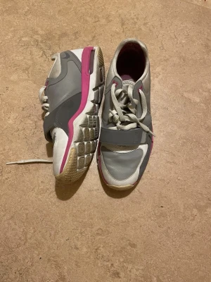 Grå och rosa sneakers med snörning - Snygga sneakers i grått och vitt med rosa detaljer. Skorna har snörning och en bred rem över foten för extra stöd. Sulan är vit med rosa inslag och har ett mönstrat grepp. Perfekta för dig som gillar sportig stil och komfort.