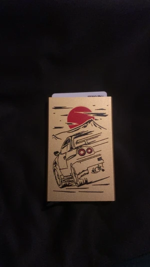 Gul metallisk plånbok med bilmotiv - Slimmad gul plånbok i metall med ett coolt tryck av en sportbil och röd sol mot berg i bakgrunden. Perfekt för kort och sedlar, lätt att bära i fickan. Snygg design som sticker ut och passar dig som gillar bilar och unik stil.