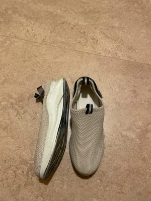 Beige slip-on sneakers med vit sula - Snygga beige slip-on sneakers med stickad ovandel och vit platt sula med svarta detaljer. Skorna har en rund tå och praktiska dragflikar både fram och bak för enkel påtagning. Perfekta för dig som gillar stilren och enkel design.