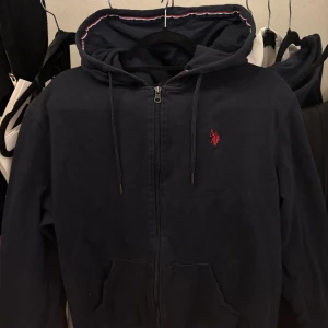 Hoddie - Mörkblå hoodie i storlek L, med dragkedja och klassisk röd logodetalj på bröstet. Bekväm, stilren och passar både vardag och träning. I fint skick och redo för en ny ägare.