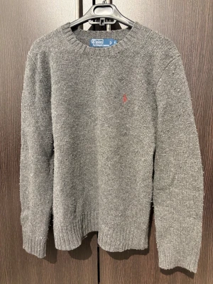 Grå stickad tröja från Polo Ralph Lauren - Snygg grå stickad tröja från Polo Ralph Lauren med klassisk rund hals och diskret röd logobroderi på bröstet. I storlek S 90% merino ull 10% angorakanninhår.