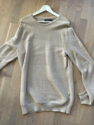 Beige partistrukturerad tröja Dressmann - Säljer en beige stickad tröja från Dressmann i storlek L. Tröjan har breda, horisontella ribbade partier som ger en cool struktur. Rund halsringning och långa ärmar. Perfekt för dig som gillar stilrena och neutrala plagg.