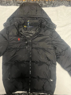 Svart pufferjacka från Polo Ralph Lauren - Svart pufferjacka från Polo Ralph Lauren med huva och två sidofickor med dragkedja. Passar 180-185 cm, Kommer med sin tag och kvitto, skriv vid någon fundering.🙌