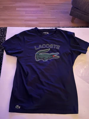 Mörkblå Lacoste t-shirt M - Snygg mörkblå t-shirt från Lacoste. Materialet är en mix av bomull och polyester som känns skönt mot huden. Säljer för att den inte kommer till användning, pris kan diskuteras vid snabb affär